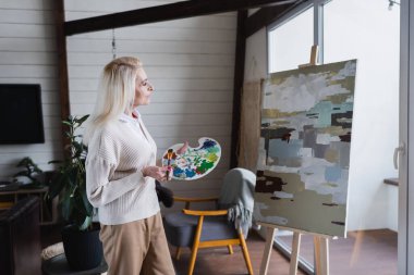 Side view of elderly woman with brush looking at painting at home 