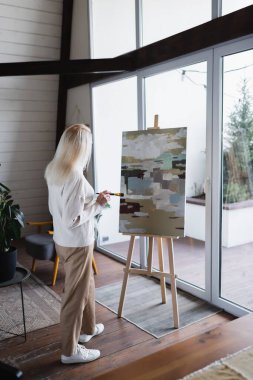 Back view of woman with paintbrush looking at canvas at home 
