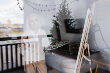 Senior woman holding palette near easel and window at home 