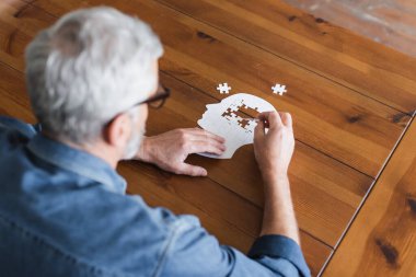 Overhead view of senior man on blurred foreground folding puzzle on table 