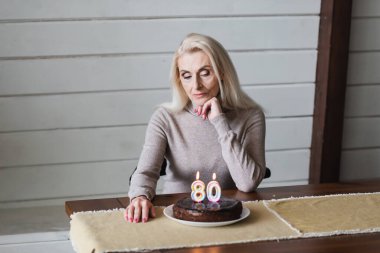 Lonely woman looking at birthday cake with candles in shape of eighty numbers 