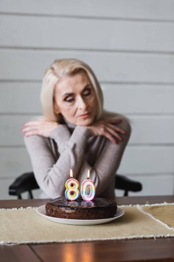 Birthday cake with candles in shape of eighty numbers on table near blurred senior woman 
