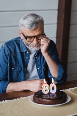 Lonely man looking at blurred birthday cake with candles in shape of eighty numbers 