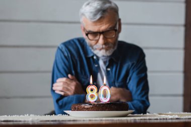 Birthday cake with burning candles in shape of eighty numbers near senior man on blurred background 