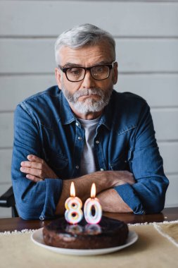 Lonely man in eyeglasses sitting near blurred birthday cake with candles in shape of eighty numbers 