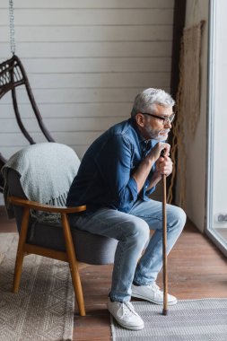 Elderly man with crutch sitting on armchair near window