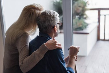 Back view of woman hugging senior husband with crutch near window 