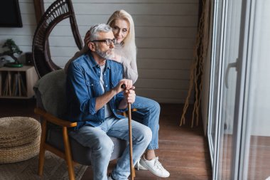 Senior man with crutch sitting on armchair near wife 