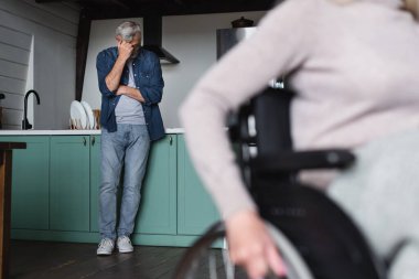 Displeased man standing in kitchen near blurred disabled wife in wheelchair