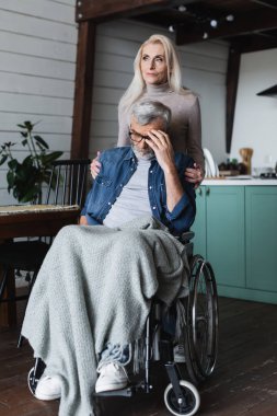 Careful woman embracing sad man in wheelchair at home  clipart