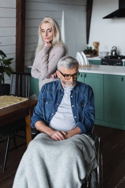 Senior woman standing near disabled husband in wheelchair 