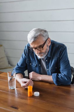 Elderly man in eyeglasses looking at pill near glass of water at home 
