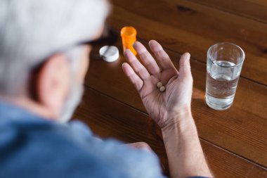 Blurred man holding pills on hand near jar and glass of water 