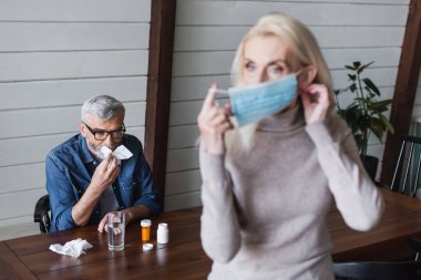 Senior man holding napkin near pills, water and wife on blurred foreground wearing medical masks 