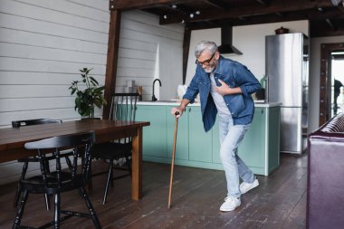 Sick senior man with crutch holding hand on chest at home 