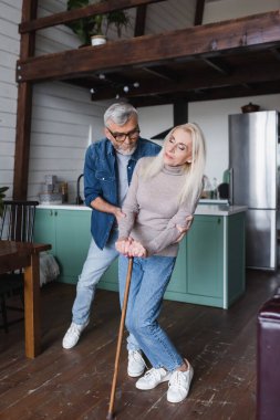 Elderly man hugging sick wife with crutch at home 