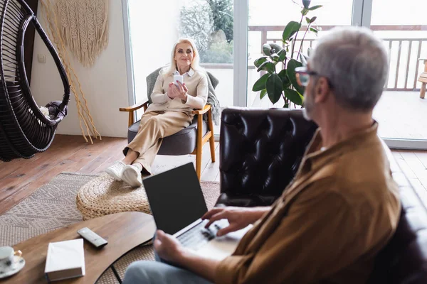 Mujer Sonriente Con Teléfono Celular Mirando Esposo Borroso Con Computadora — Foto de Stock