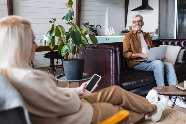 在客厅用笔记本电脑微笑的老年男子接近模糊的妻子和手机 — 图库照片