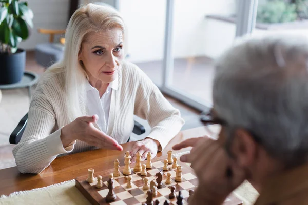 Femme Âgée Pointant Vers Les Échecs Près Mari Flou Maison — Photo