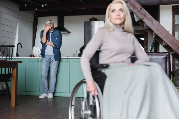 Triste Uomo Piedi Cucina Vicino Moglie Offuscata Sedia Rotelle — Foto Stock