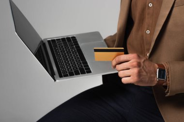 Cropped view of businessman in brown jacket holding credit card and blurred laptop isolated on grey  clipart