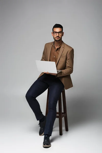 Arabian Businessman Looking Camera While Holding Laptop Chair Grey Background — Zdjęcie stockowe