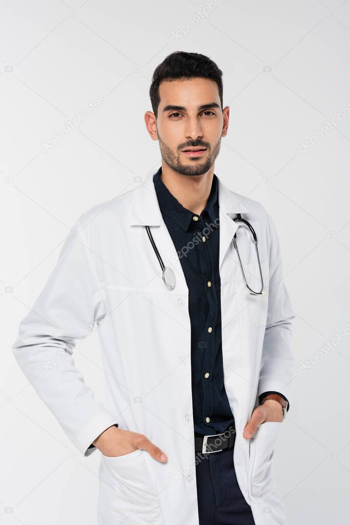 Arabian doctor with stethoscope holding hands in pockets of white coat isolated on grey 