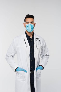 Tıbbi maskeli ve lateks eldivenli Arap doktor gri kameraya bakıyor. 