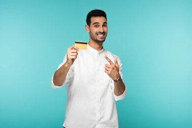 Cheerful arabian man pointing at credit card isolated on blue  clipart