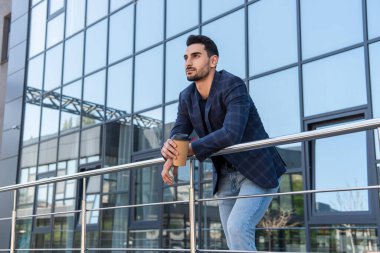 Young muslim businessman with takeaway drink looking away near railing outdoors  clipart