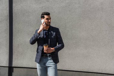 Elinde kağıt bardak tutan ve beton duvarın yanında akıllı telefondan konuşan Müslüman bir adam. 