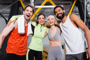 Spor salonunda kucaklaşırken kameraya bakan neşeli çoklu etnik insanlar