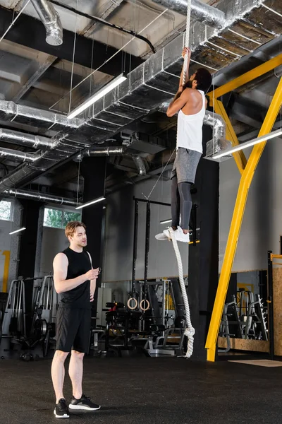 Trainer Looking Fitness Tracker African American Man Climbing Rope — Stock Photo, Image