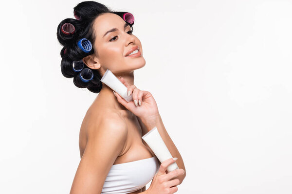 улыбающаяся молодая женщина с бигуди на голове держа кремовые трубки возле шеи и груди изолированы на белом