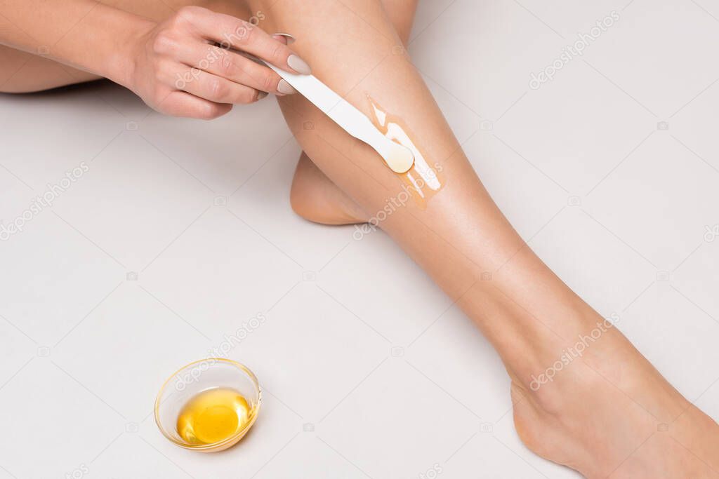 close up view of young woman making epilation on leg near bowl with honey on white