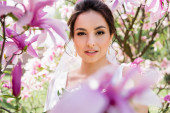Bruneta nevěsta při pohledu na kameru v blízkosti rozmazané magnólie květiny 