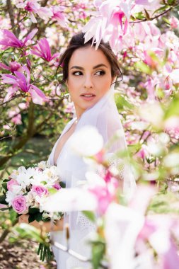 Bride with bouquet looking away near blooming magnolia tree  clipart
