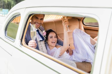 Neşeli damat, eski arabanın içinde, gelinin yanında bulanık bir kadeh şampanya tutuyor. 