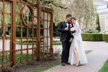 Gelin elbisesi ve parkta çiçek açan ağacın yanında duran neşeli damat. 