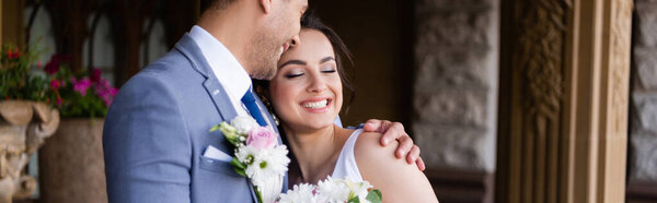 Cheerful groom hugging bride with closed eyes outdoors, banner 