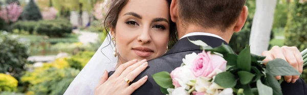 Fröhliche Braut Umarmt Bräutigam Und Hält Blumenstrauß Auf Verschwommenem Vordergrund — Stockfoto