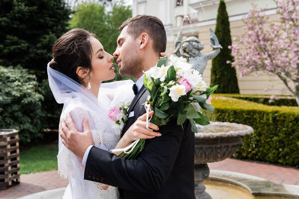 Side view of newlyweds with bouquet kissing near fountain 