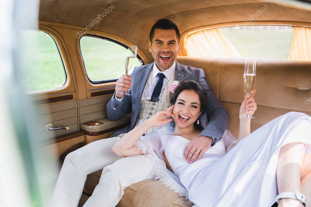 Happy newlyweds with glasses of champagne looking at camera in car 