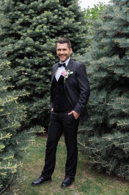 Gülümseyen damat parkta çam ağaçlarının yanında pantolon ceplerinde el ele tutuşuyor. 