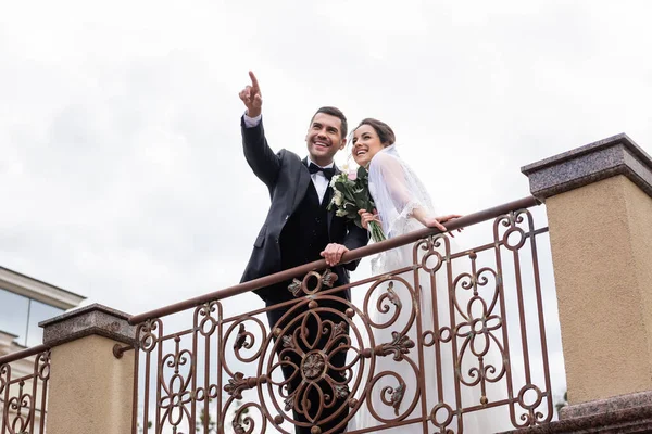 Low angle view of positive groom pointing with finger near cheerful bride on bridge