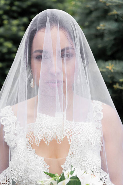 Bride in veil looking at camera near flowers on blurred foreground outdoors 