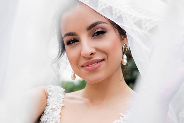 Smiling bride looking at camera near blurred white veil 