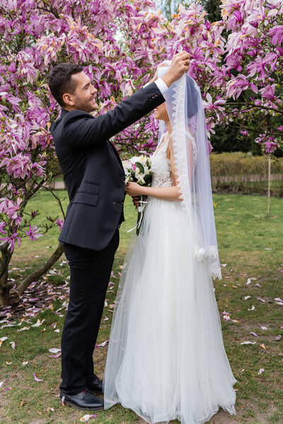 Вид сбоку улыбающегося жениха, снимающего вуаль с невесты возле деревьев магнолии 