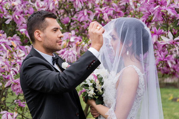 Вид сбоку на жениха, снимающего вуаль с невесты с букетом в парке 