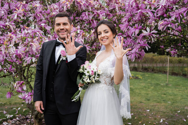 Улыбающиеся невеста и жених показывают кольца возле деревьев магнолии 